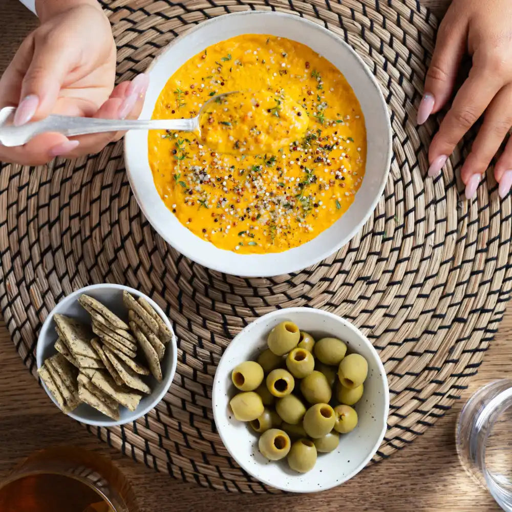 A bowl of creamy orange dip garnished with herbs and spices, served with crackers and green olives.