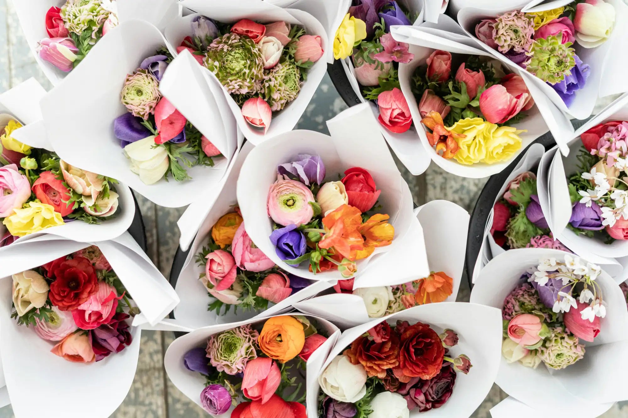 A vibrant bouquet of assorted ranunculus and other flowers in white paper wraps, featuring a mix of pink, purple, yellow, orange, red, and cream blooms.